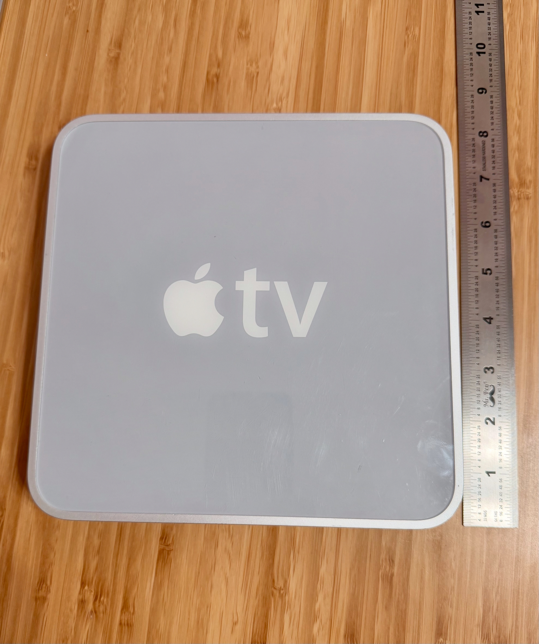 A square Apple TV device is placed on a wooden surface next to a ruler.