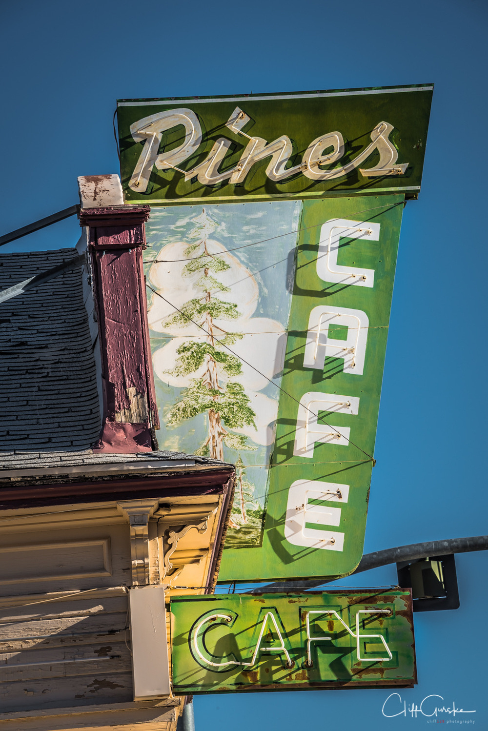 An old, weathered sign for Pines Cafe with a pine tree illustration is set against a clear blue sky.