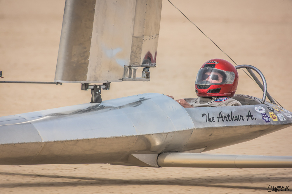 A person wearing a red helmet sits in a streamlined, metallic land yacht with The Arthur A written on the side.