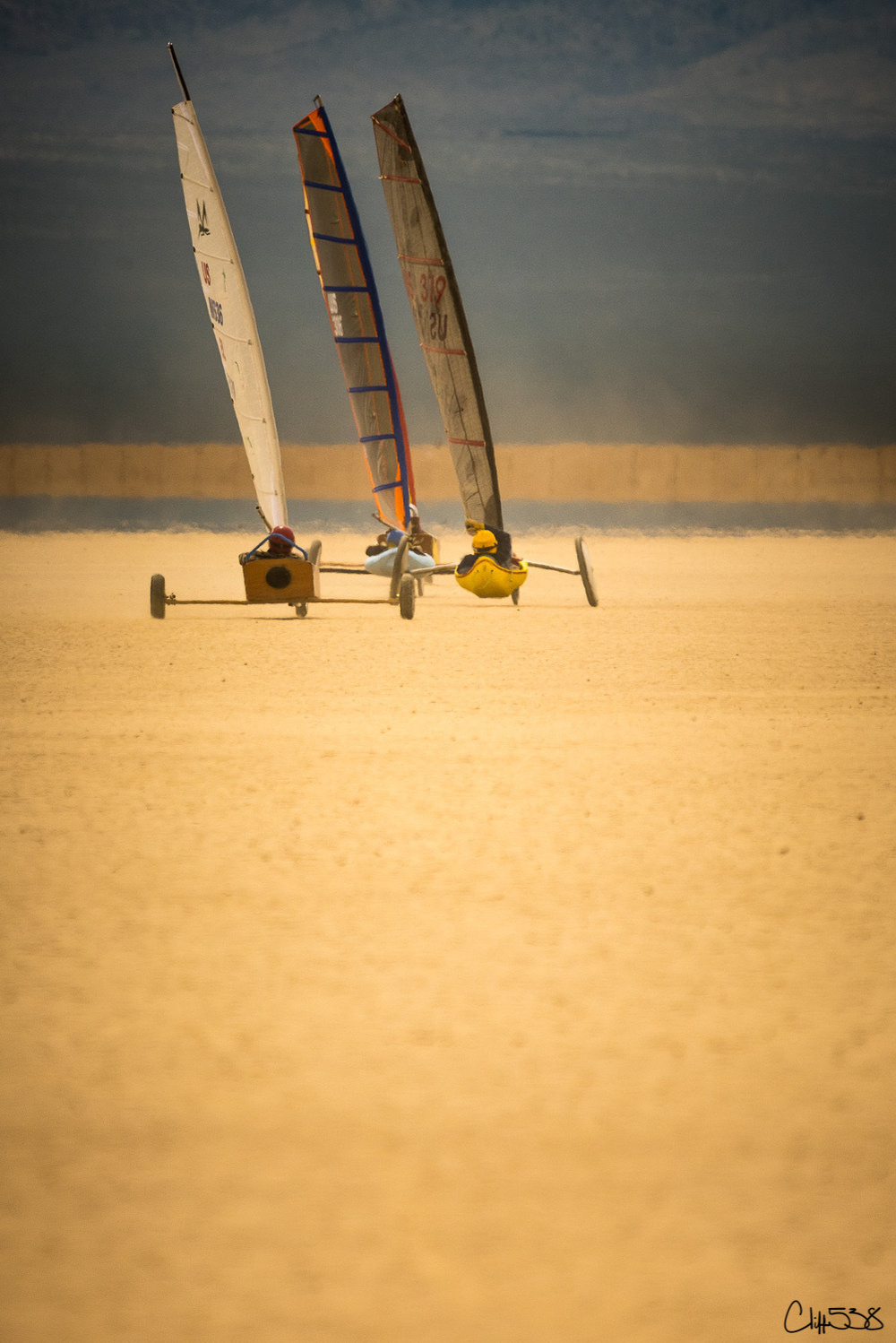 Three land yachts race across a vast, sandy terrain under a cloudy sky.