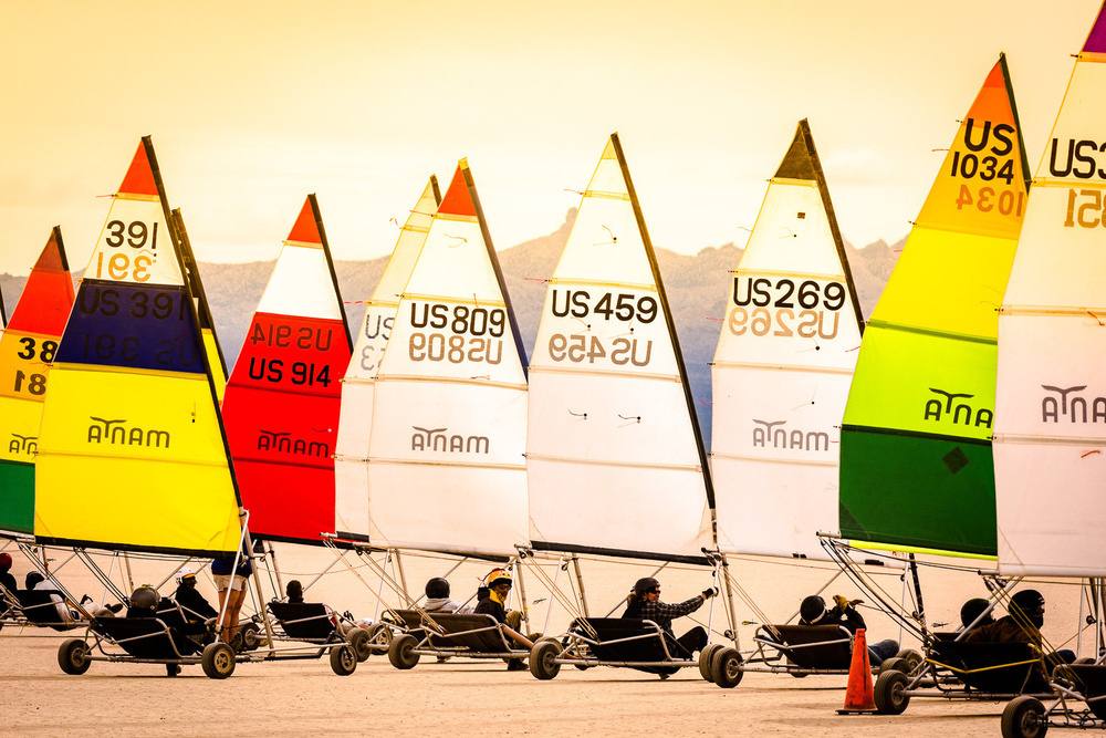 Several colorful land sailboats with their sails marked by identification numbers are lined up on a desert landscape.