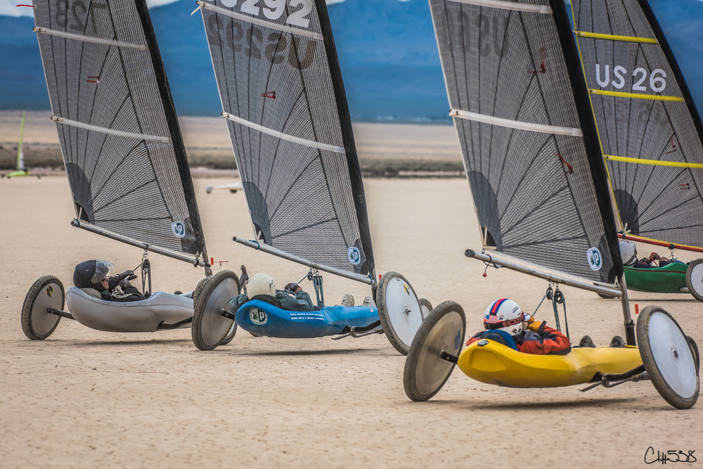 Three people are racing on land yachts across a dry, sandy terrain with mountains in the background.