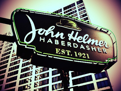 A vintage-style sign for John Helmer Haberdasher, Est. 1921 is displayed against a backdrop of tall buildings.