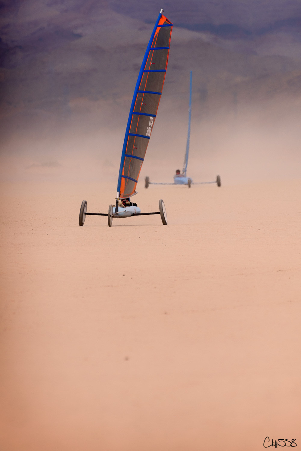 Two land sailboats race across a vast, sandy landscape with a hazy mountain backdrop.