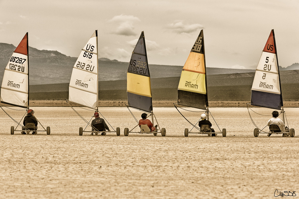 Several land yachts with colorful sails are lined up on a dry desert plain, ready for racing.