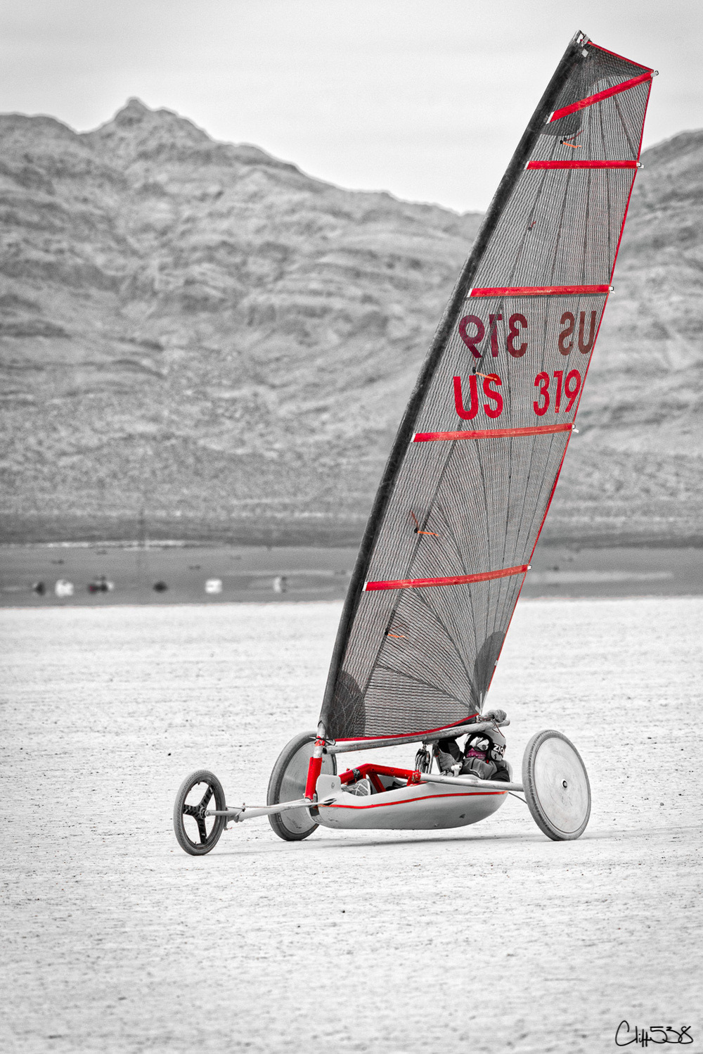 A land yacht with a large sail is being piloted on a dry, flat terrain with mountains in the background.