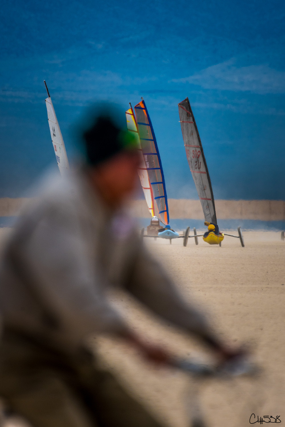 A person on a bicycle appears out of focus in the foreground while three land sailboats with colorful sails are seen on a beach in the background.