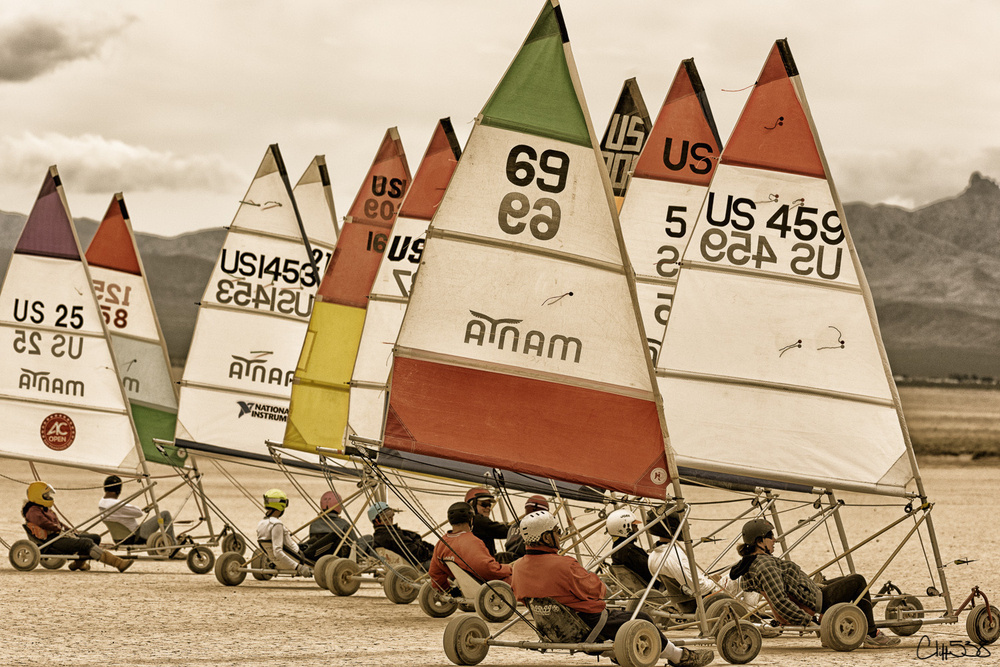 Several colorful land sailboats with numbered sails are lined up on a dry desert lakebed, each piloted by individuals wearing helmets.