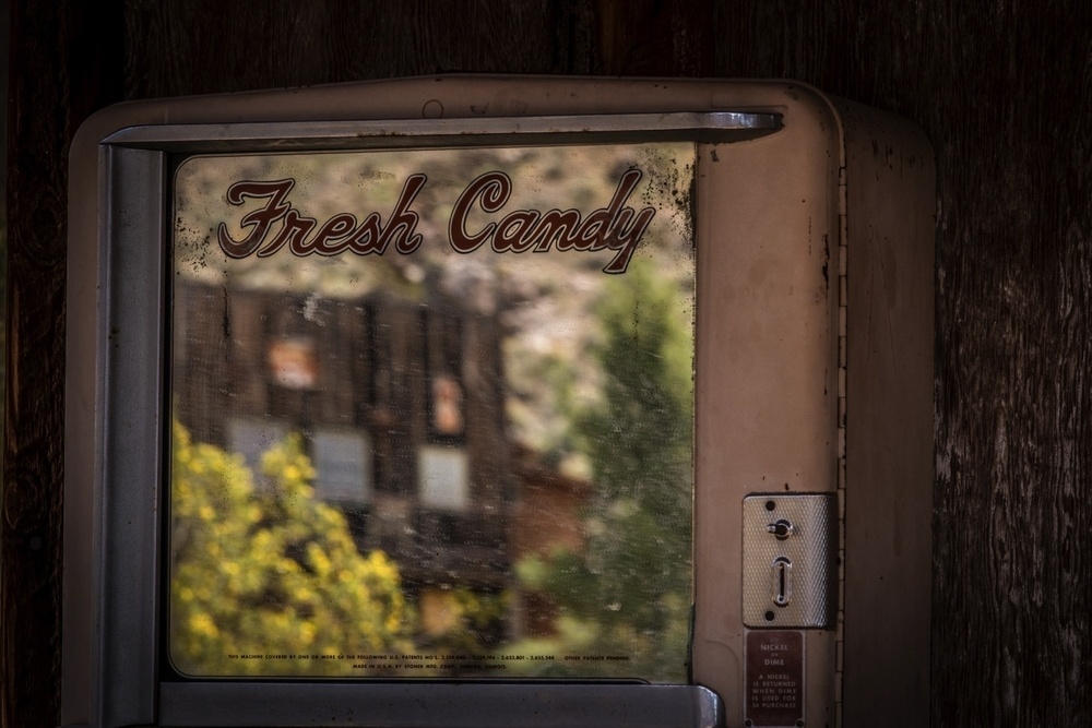 A vintage candy vending machine with the text Fresh Candy is positioned in front of a blurred building and trees in the background.