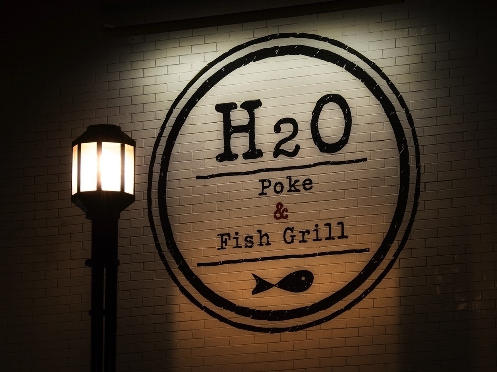 A round sign for H2O Poke & Fish Grill is illuminated on a brick wall next to a street lamp.