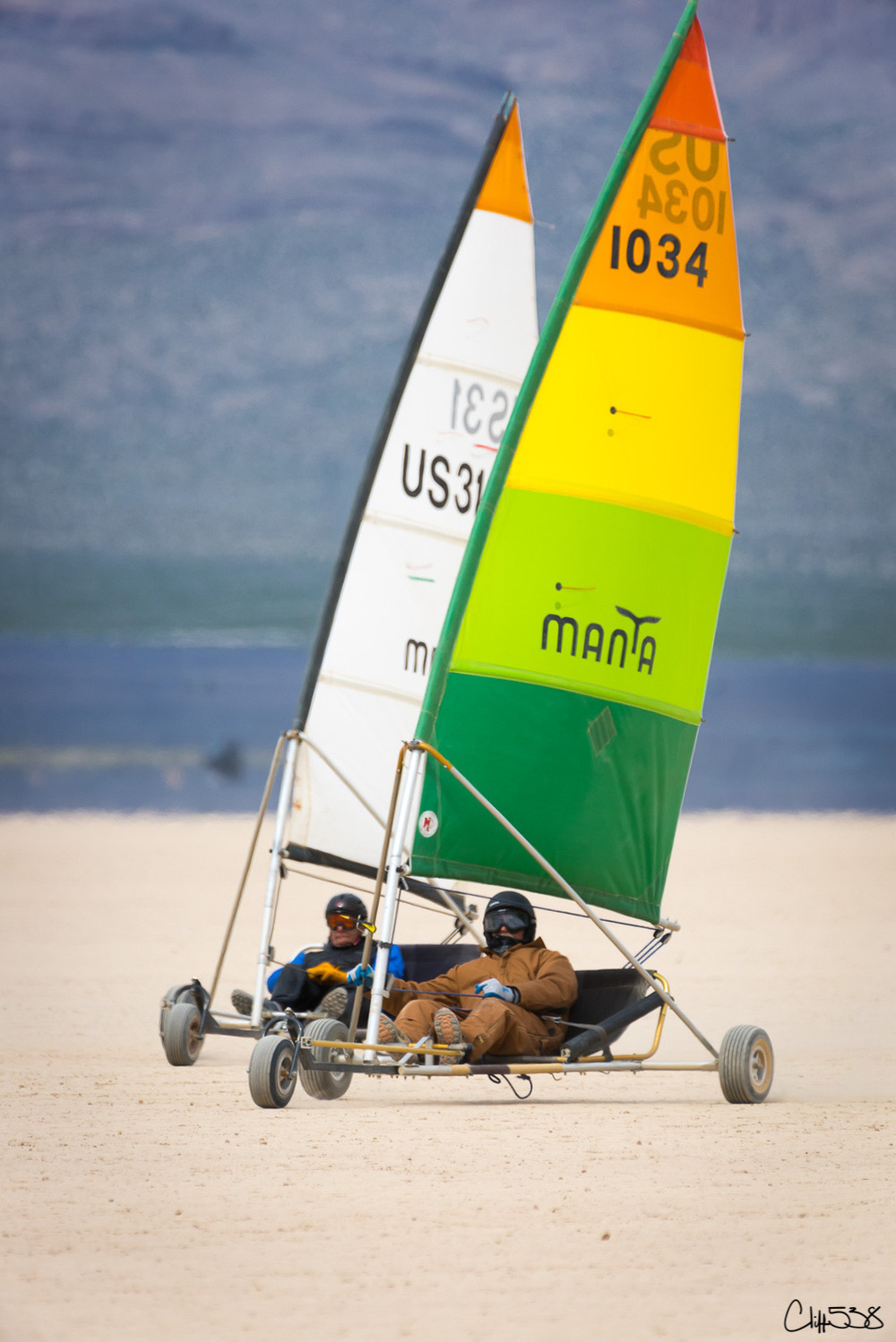 Two people are seated in colorful land sailboats racing on a sandy terrain.