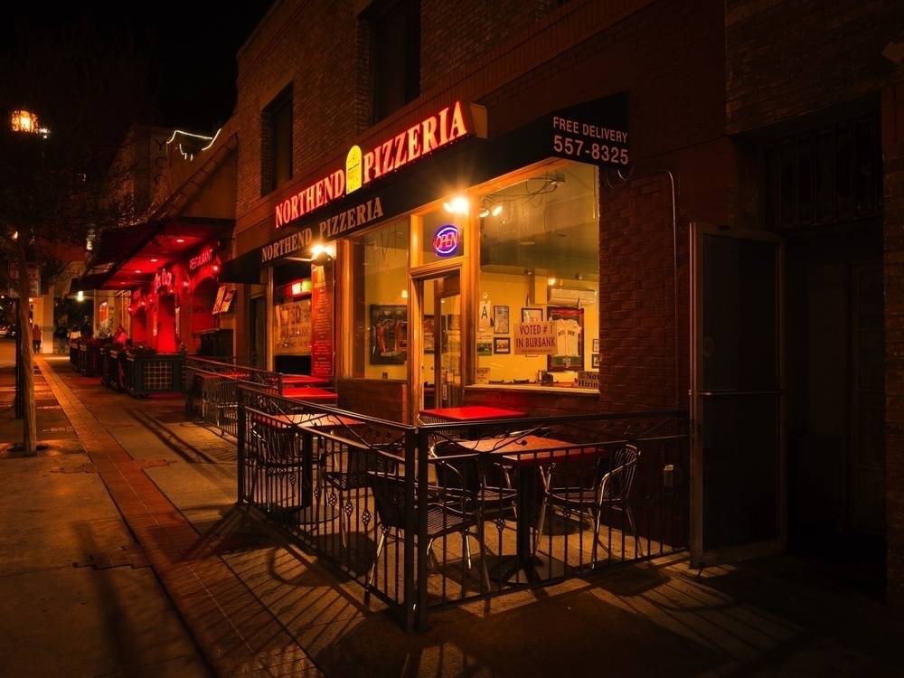 A dimly lit pizzeria with outdoor seating is visible at night, featuring a neon open sign and illuminated interior.