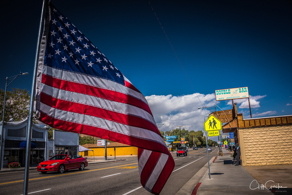 An American flag flutters in the foreground beside a road with buildings, a red car, and a pedestrian crossing sign under a clear blue sky.