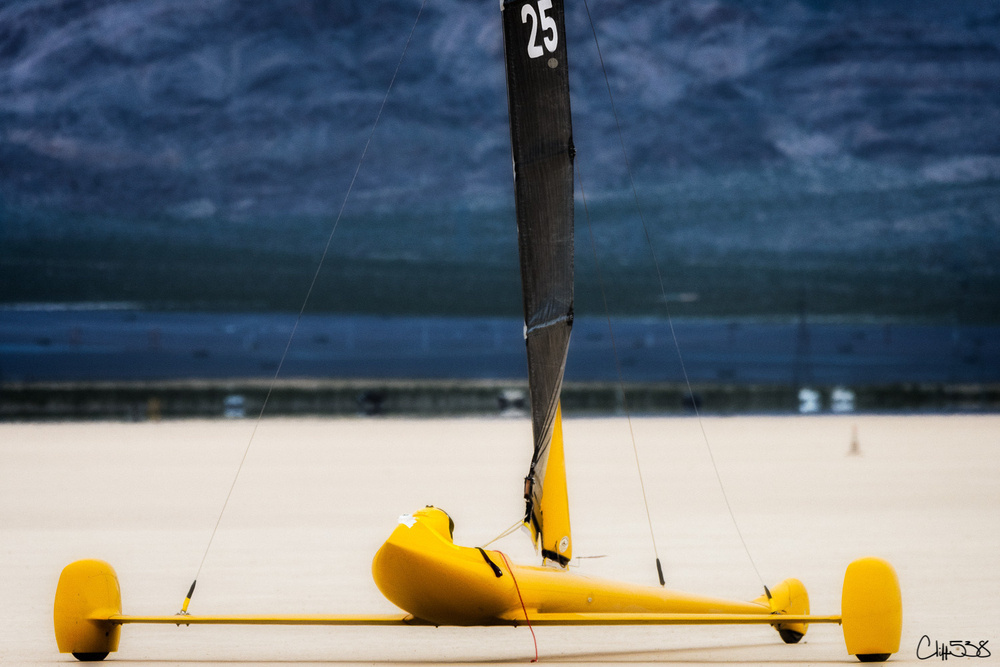 A yellow land yacht with a black sail is on a vast, flat desert landscape.