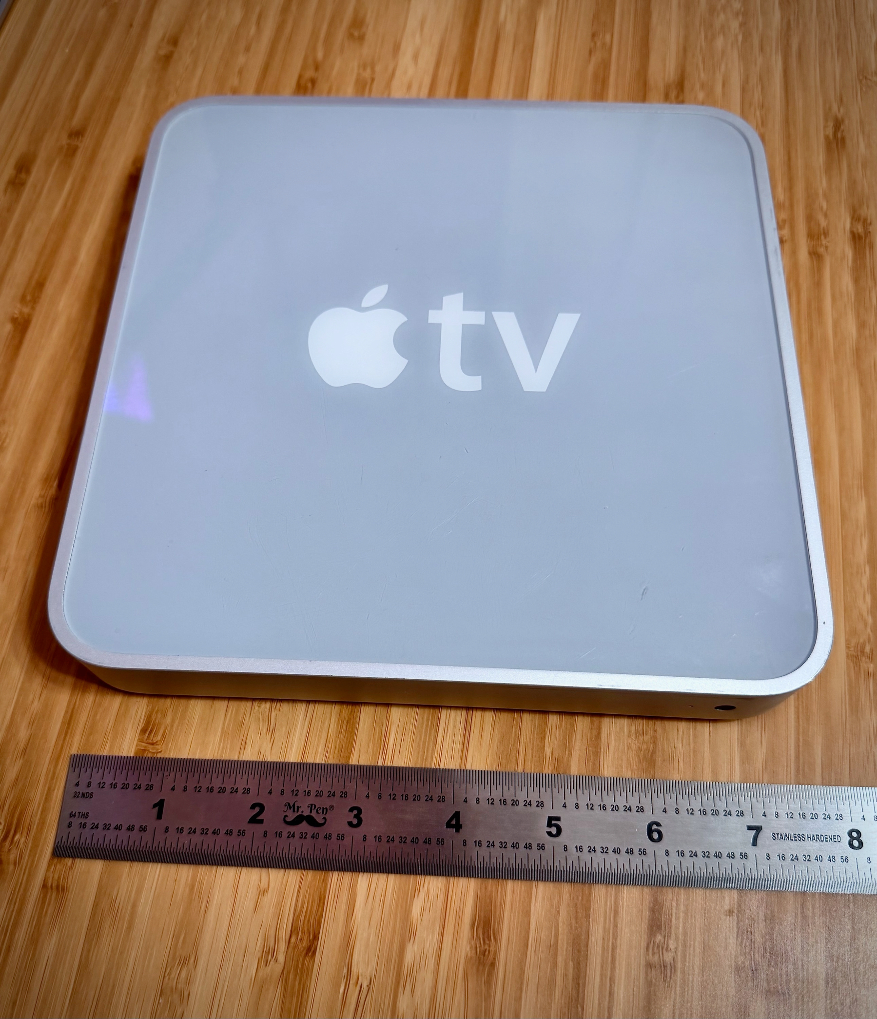 A silver Apple TV device is placed on a wooden surface next to a ruler.