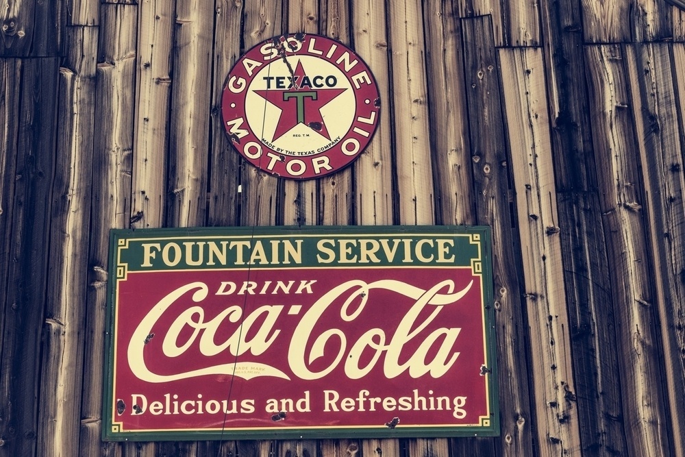 A vintage Texaco gasoline sign and a retro Coca-Cola advertisement are displayed on a wooden wall.