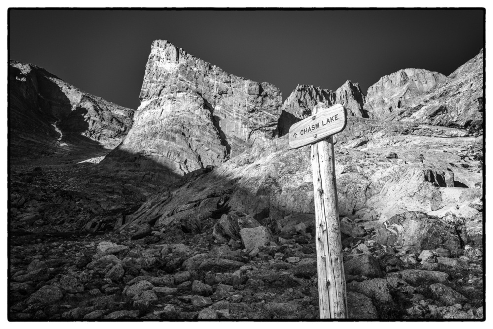 A rugged mountain landscape features a wooden signpost labeled CHASM LAKE in the foreground.