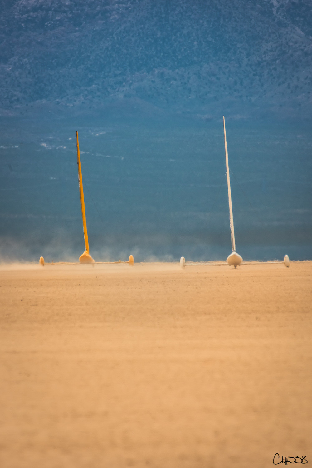 Two sailboats race across a vast, dry desert landscape with mountains in the background.