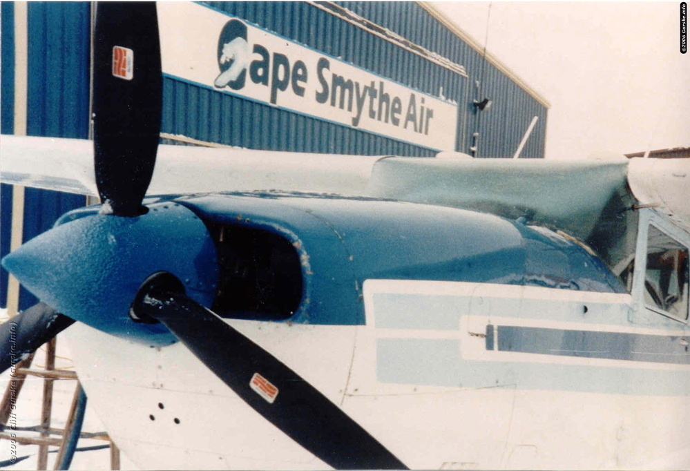 A small blue and white propeller airplane is parked in front of a building with a sign reading Cape Smythe Air.