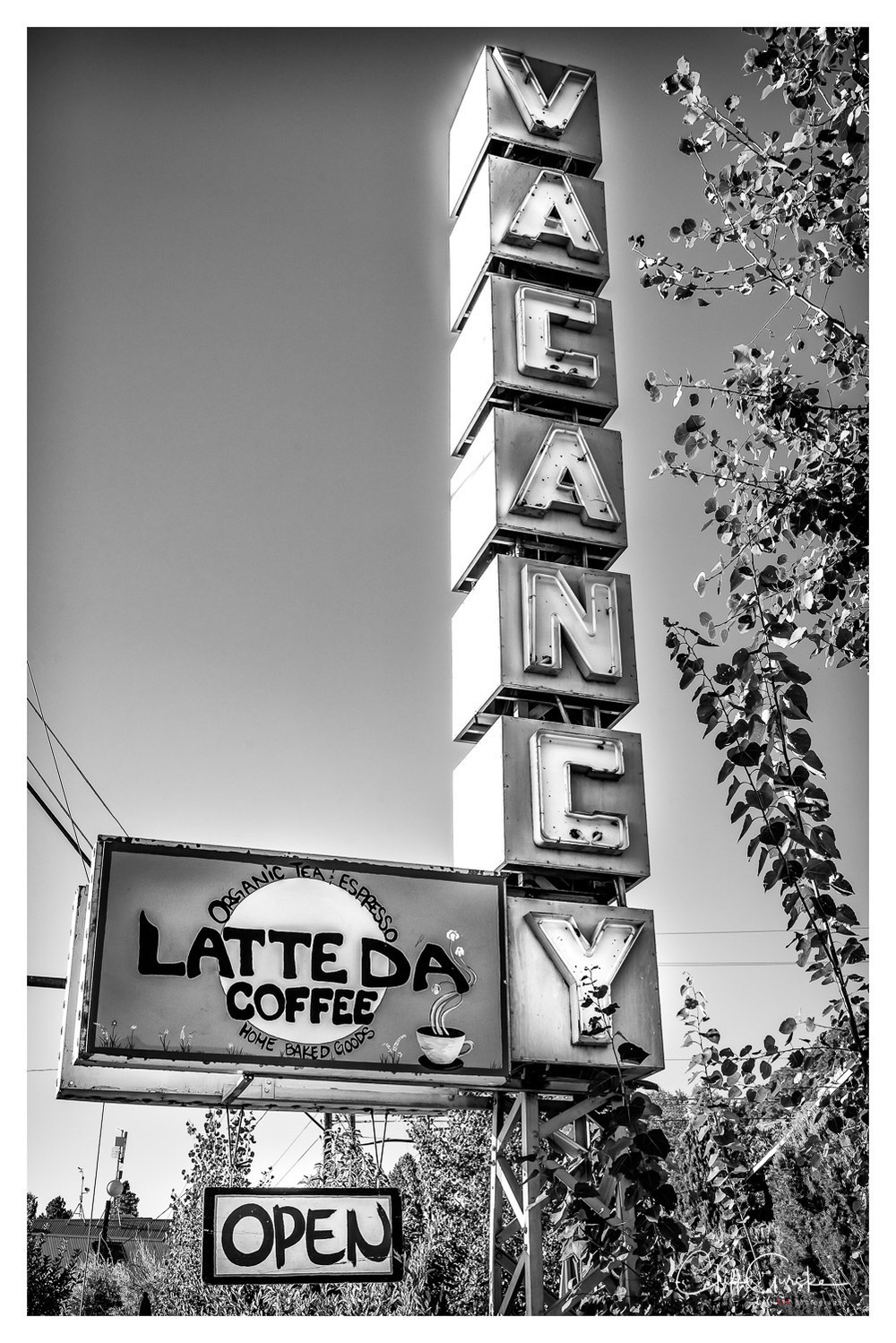 A vintage-style sign displays VACANCY vertically and LATTE DA COFFEE with the word OPEN at the bottom, set against a backdrop of trees and clear sky.