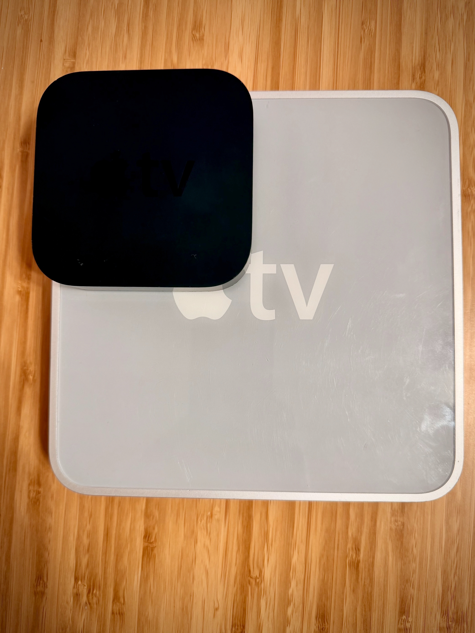 A black Apple TV device is placed on top of its large square box with a visible logo on a wooden surface.