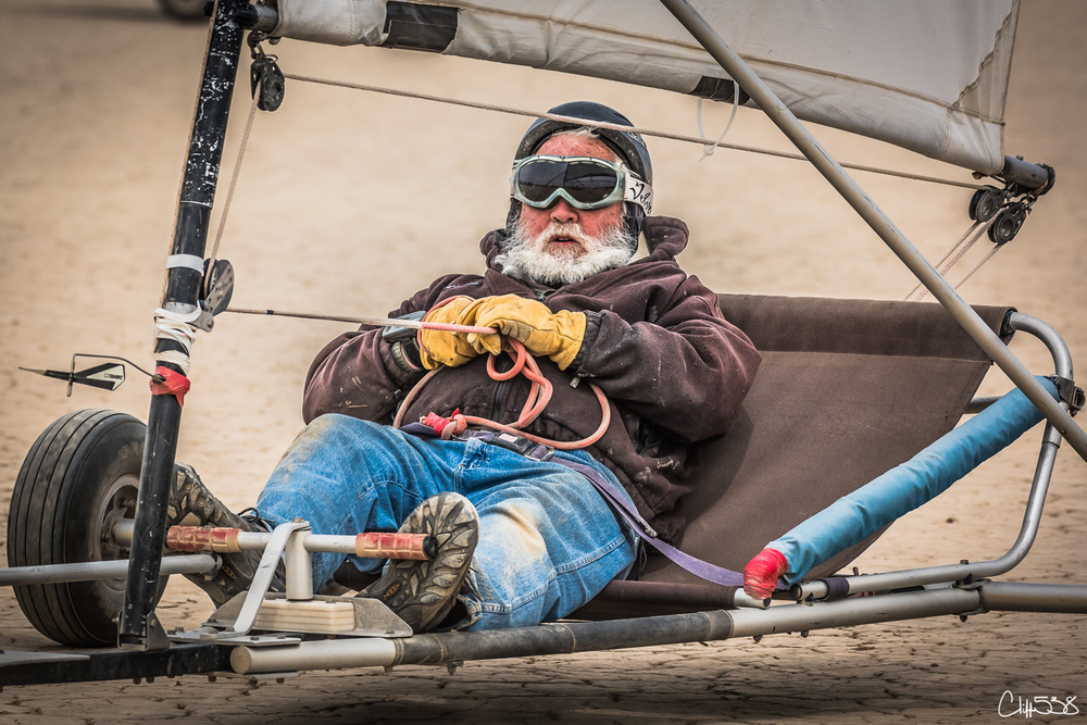 A person is sitting in a land sailer wearing goggles and gloves, ready for sailing on a sandy surface.