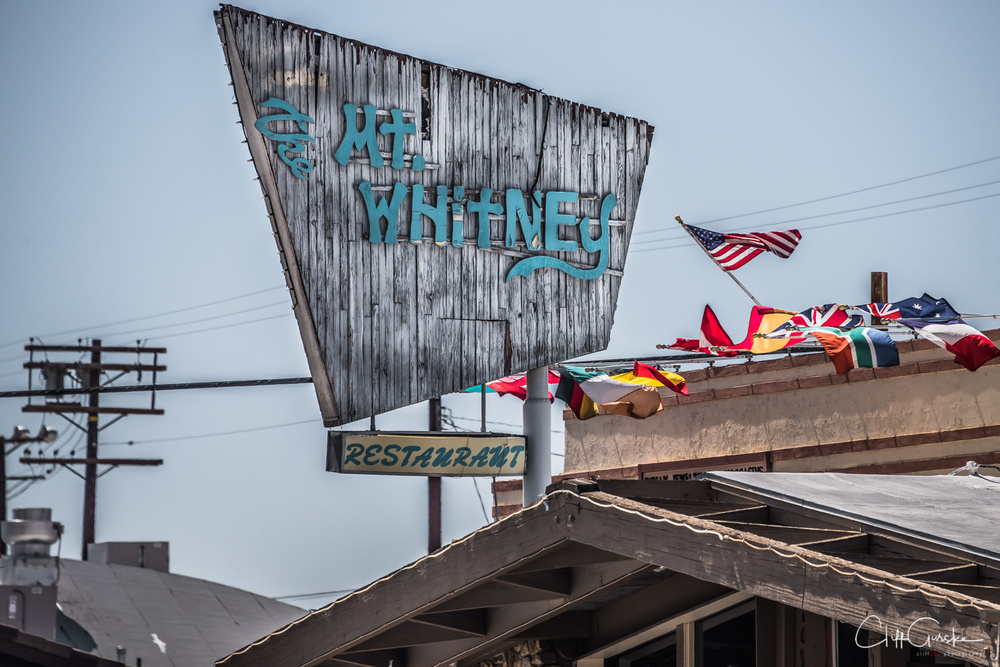 A rustic restaurant sign reads Mt. Whitney with various flags displayed on the building's rooftop.