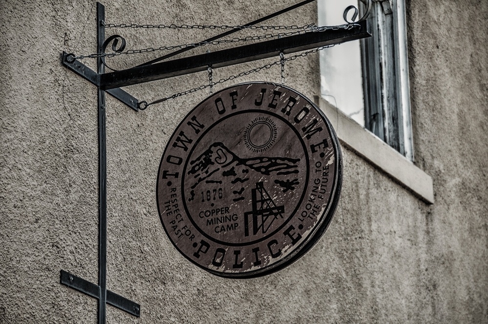 A circular sign for the Town of Jerome Police, featuring engravings related to copper mining and a rustic design, hangs on a textured wall.