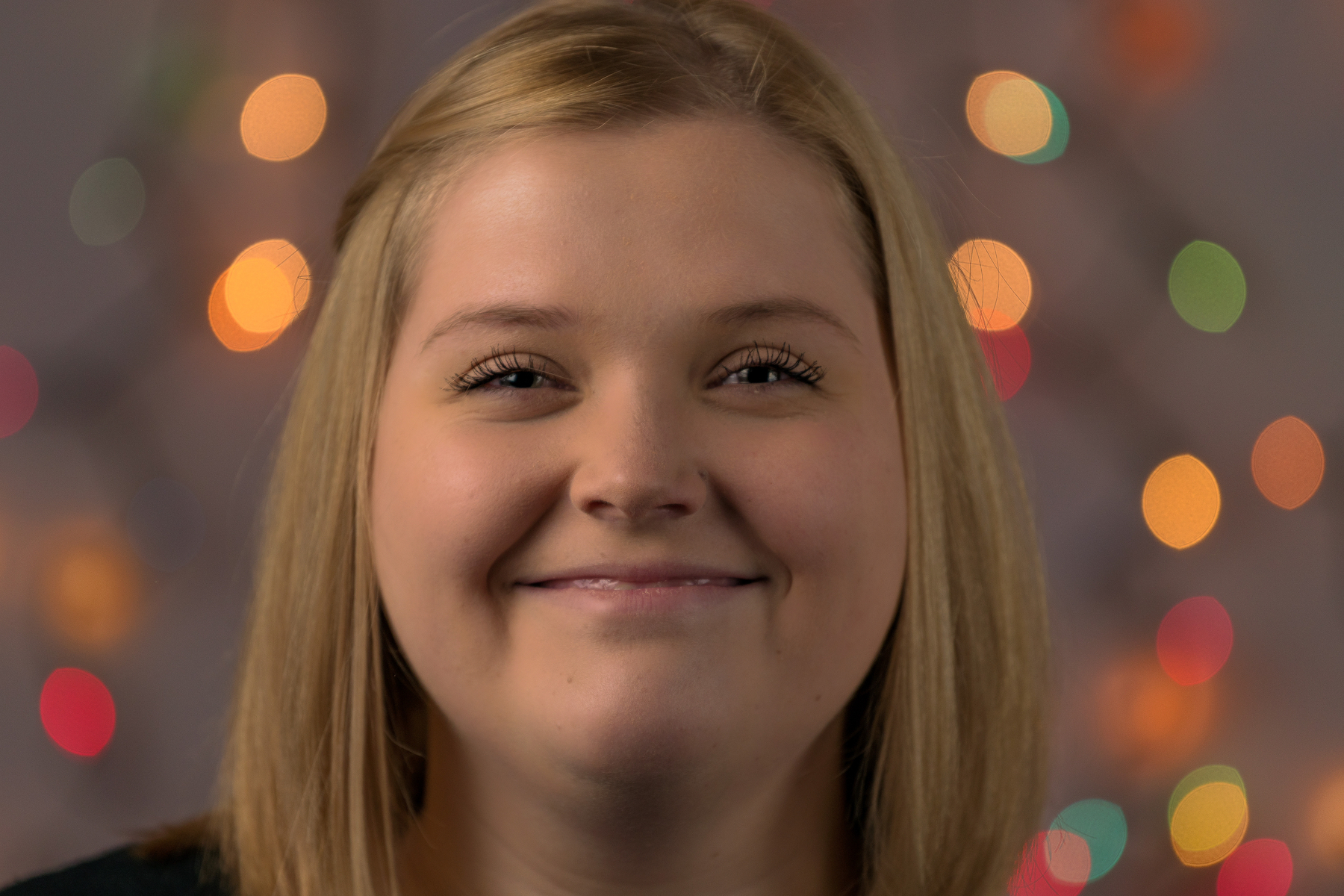 Auto-generated description: A person with blond hair is smiling in front of a backdrop with colorful, out-of-focus lights.