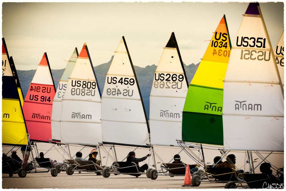 Several colorful land sailboats are lined up on a sandy terrain with mountains in the background.