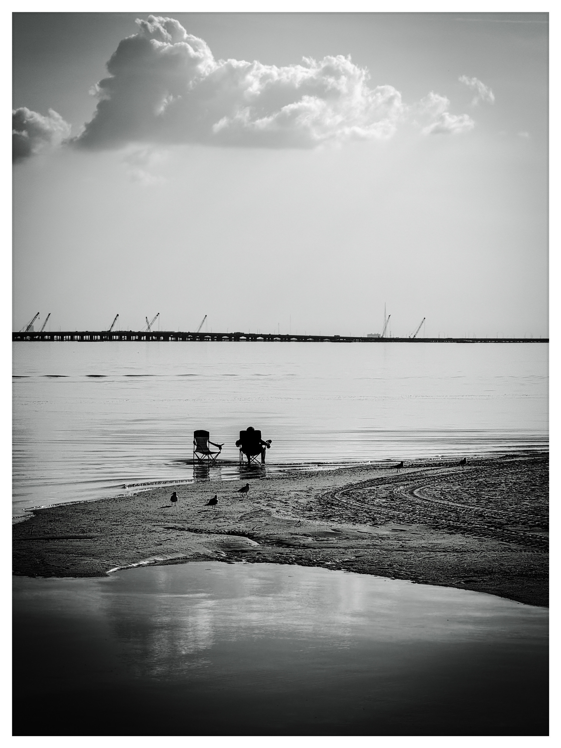 Two foldable chairs face a calm body of water with distant cranes on the horizon and a cloudy sky above.