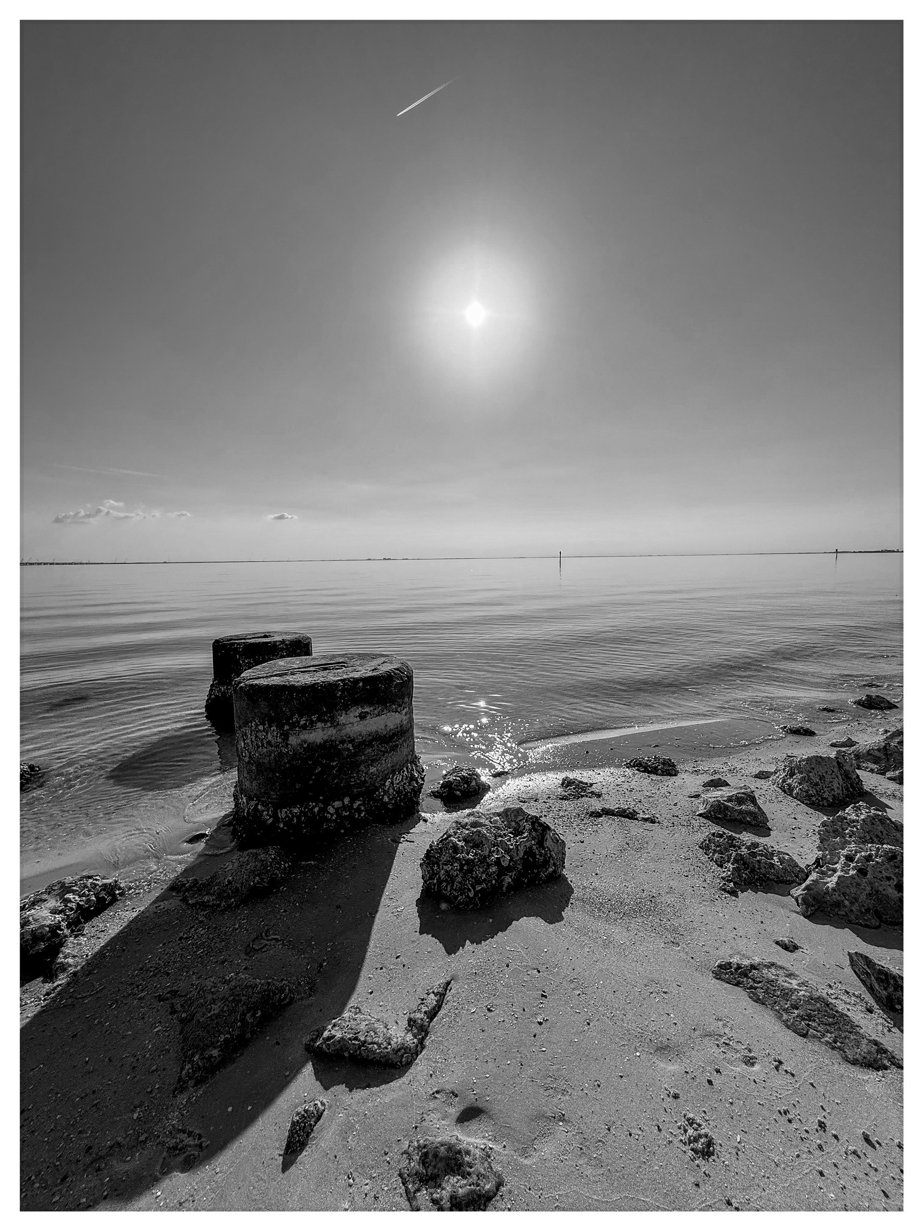 A tranquil black and white scene of a sunny beach with rocks in the foreground and a vast open ocean extending to the horizon under a clear sky.