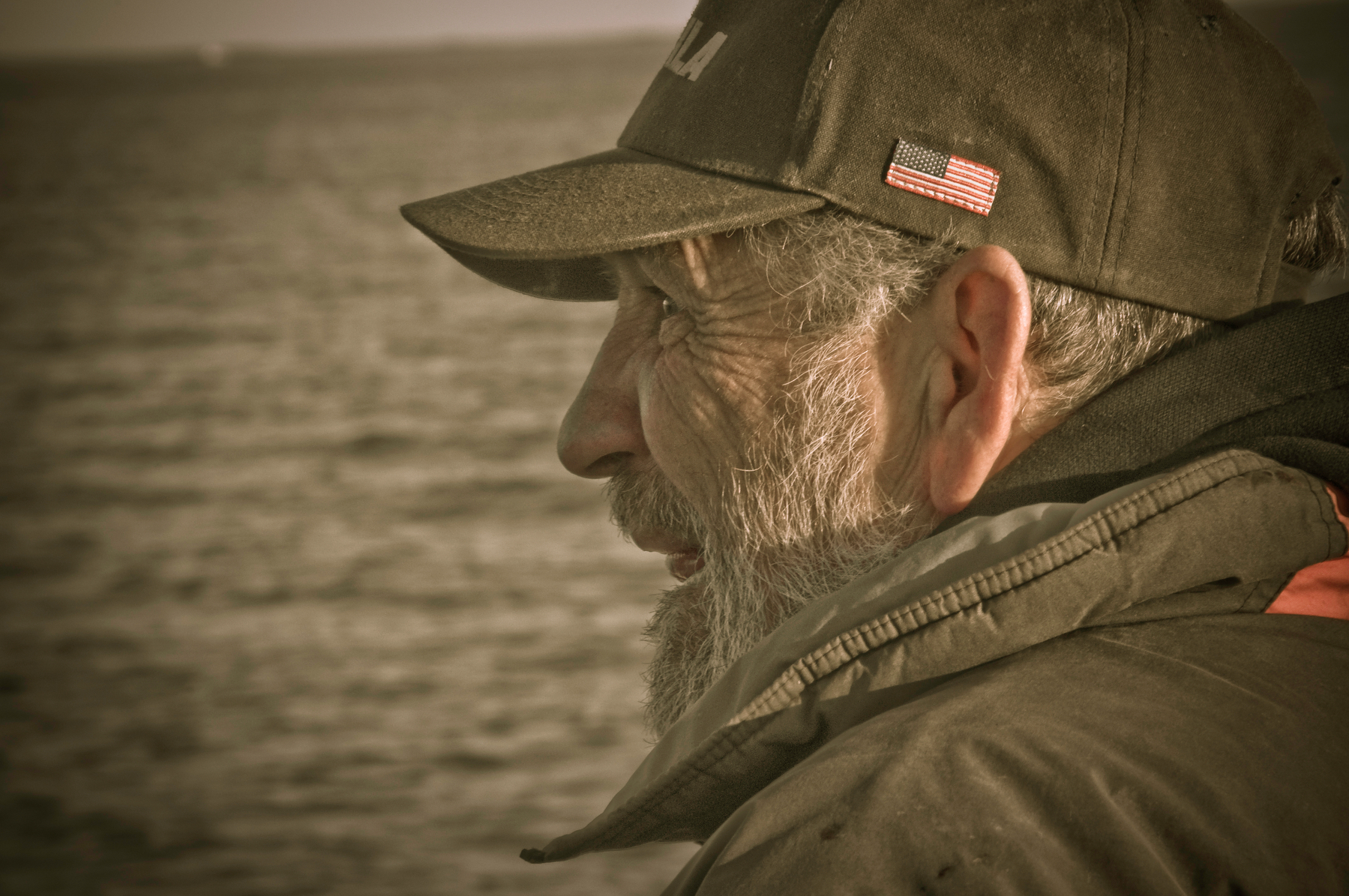 Auto-generated description: An older man wearing a hat with an American flag is gazing towards the water.