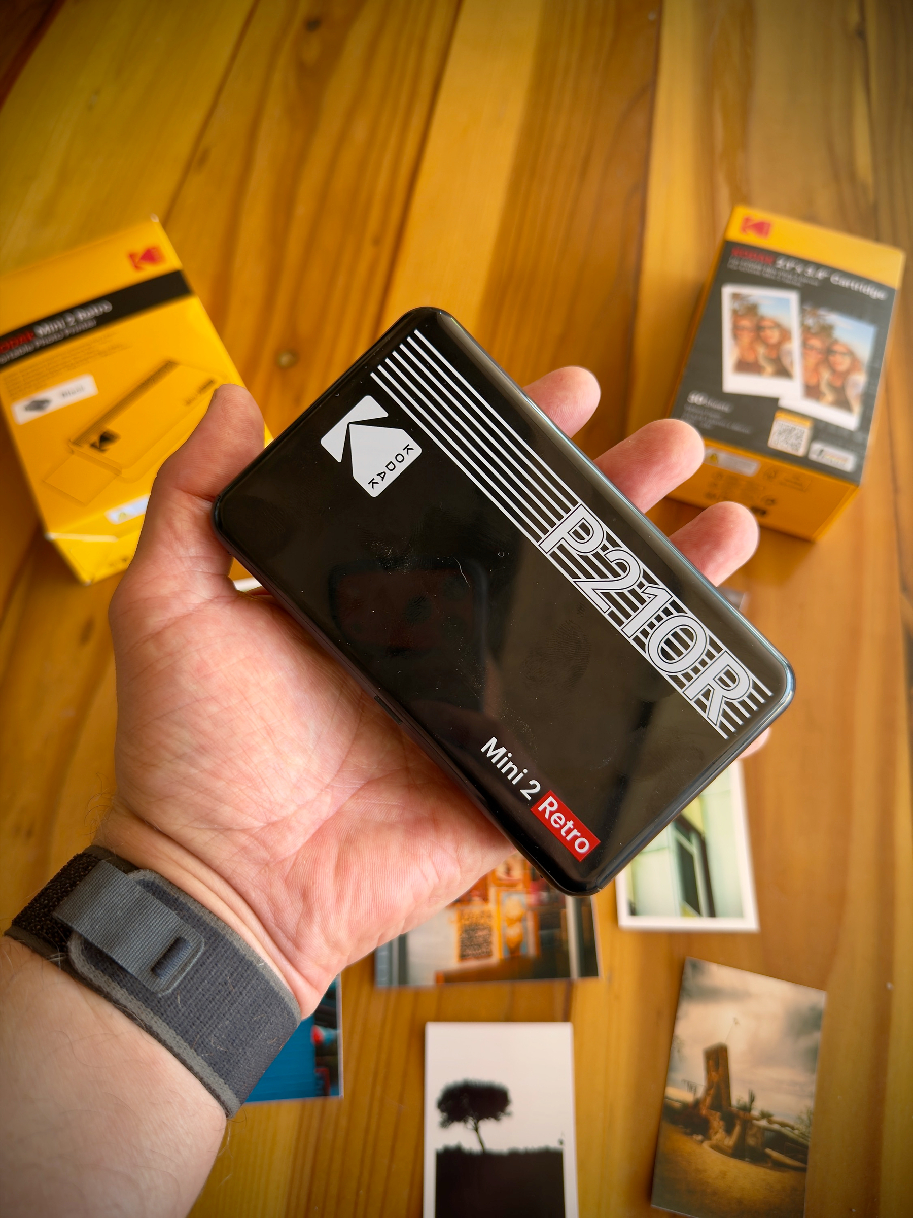 Auto-generated description: A hand is holding a Kodak P210R Mini 2 Retro photo printer, with Kodak packaging and printed photos visible in the background on a wooden surface.
