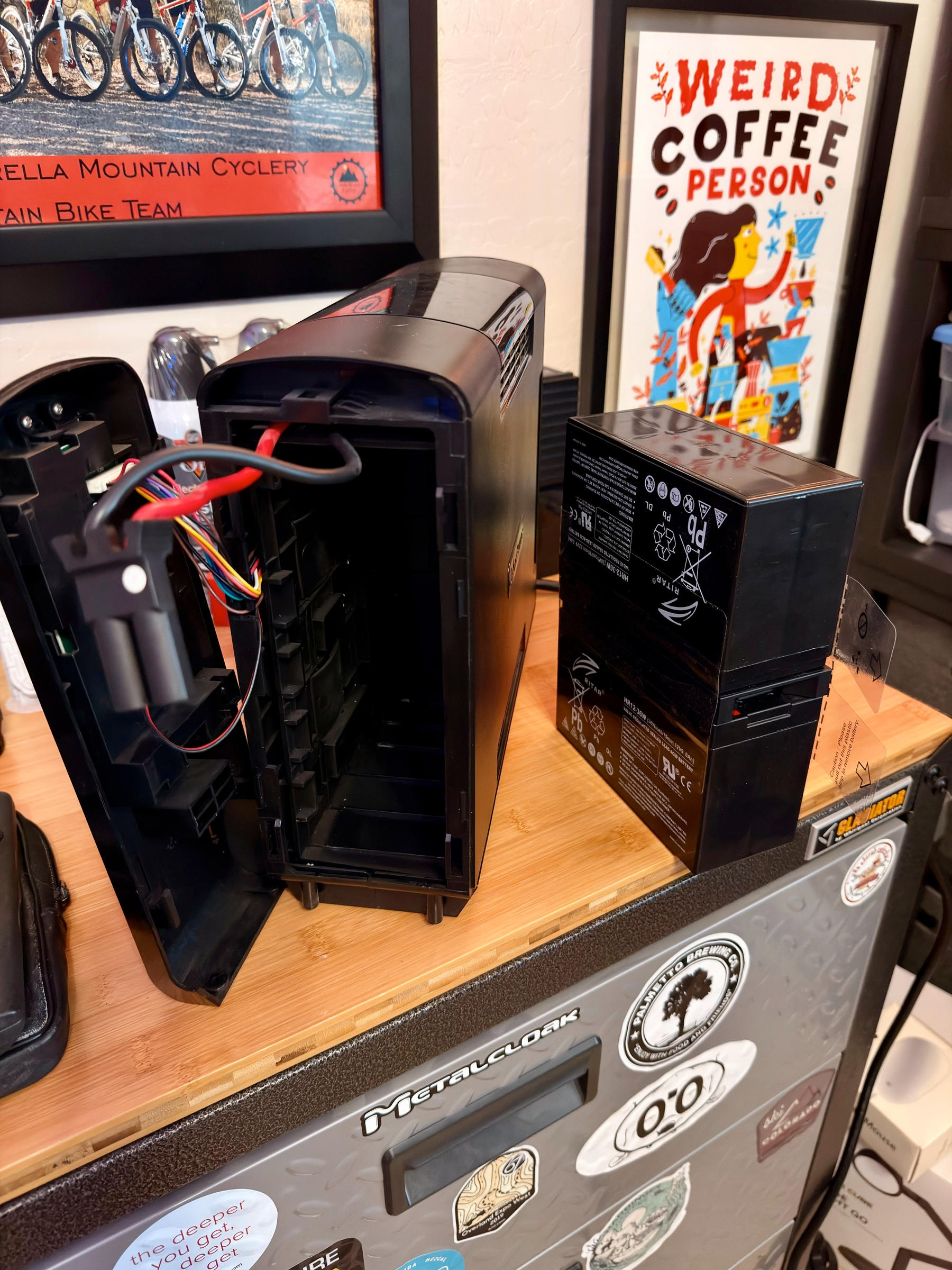Auto-generated description: A disassembled computer case sits on a desk alongside its power supply unit, with posters and bicycle-related items visible in the background.