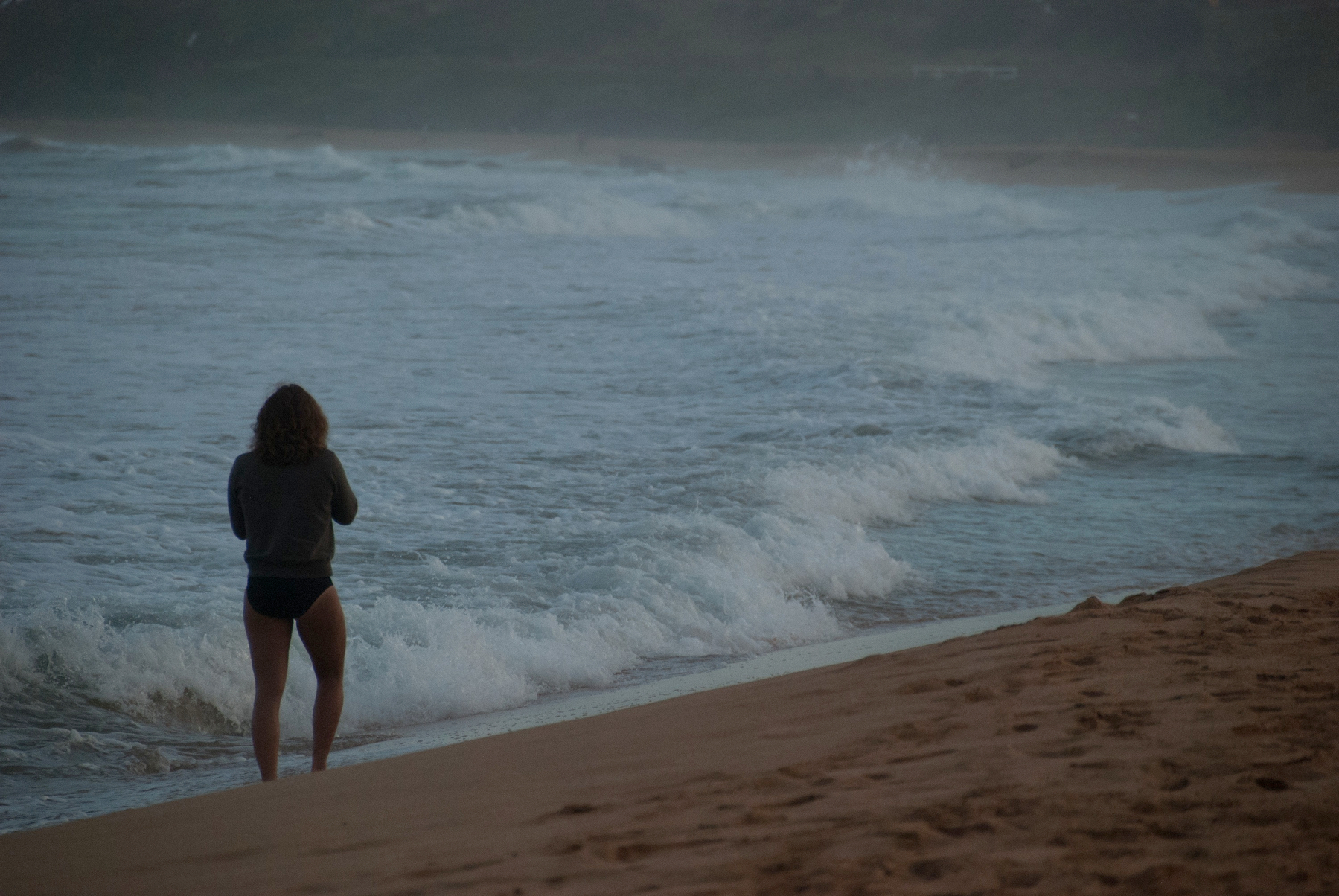 Auto-generated description: A person stands on a sandy beach facing the ocean waves.