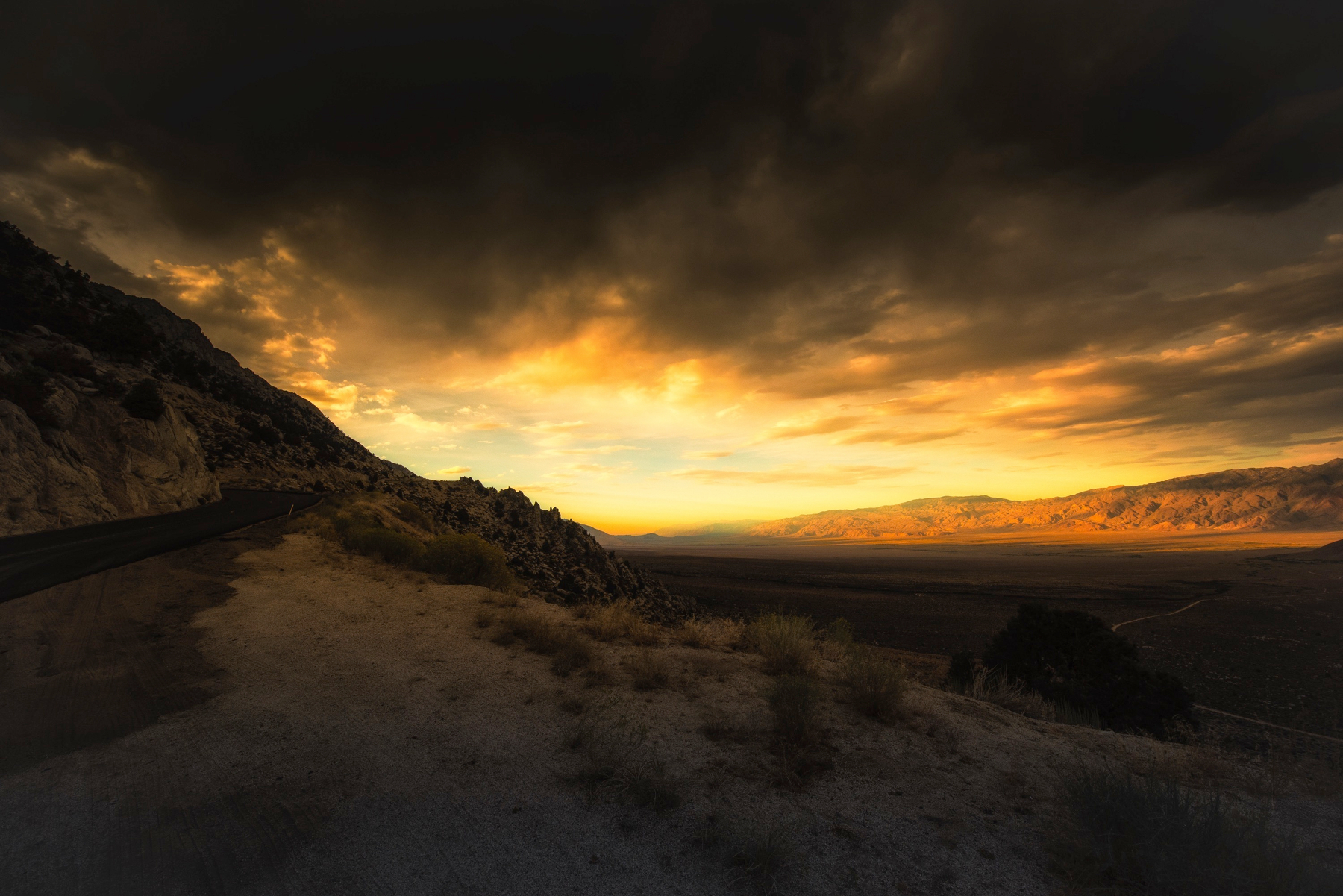 Auto-generated description: A dramatic sunset casts golden light over a rugged mountainous landscape with a winding road.