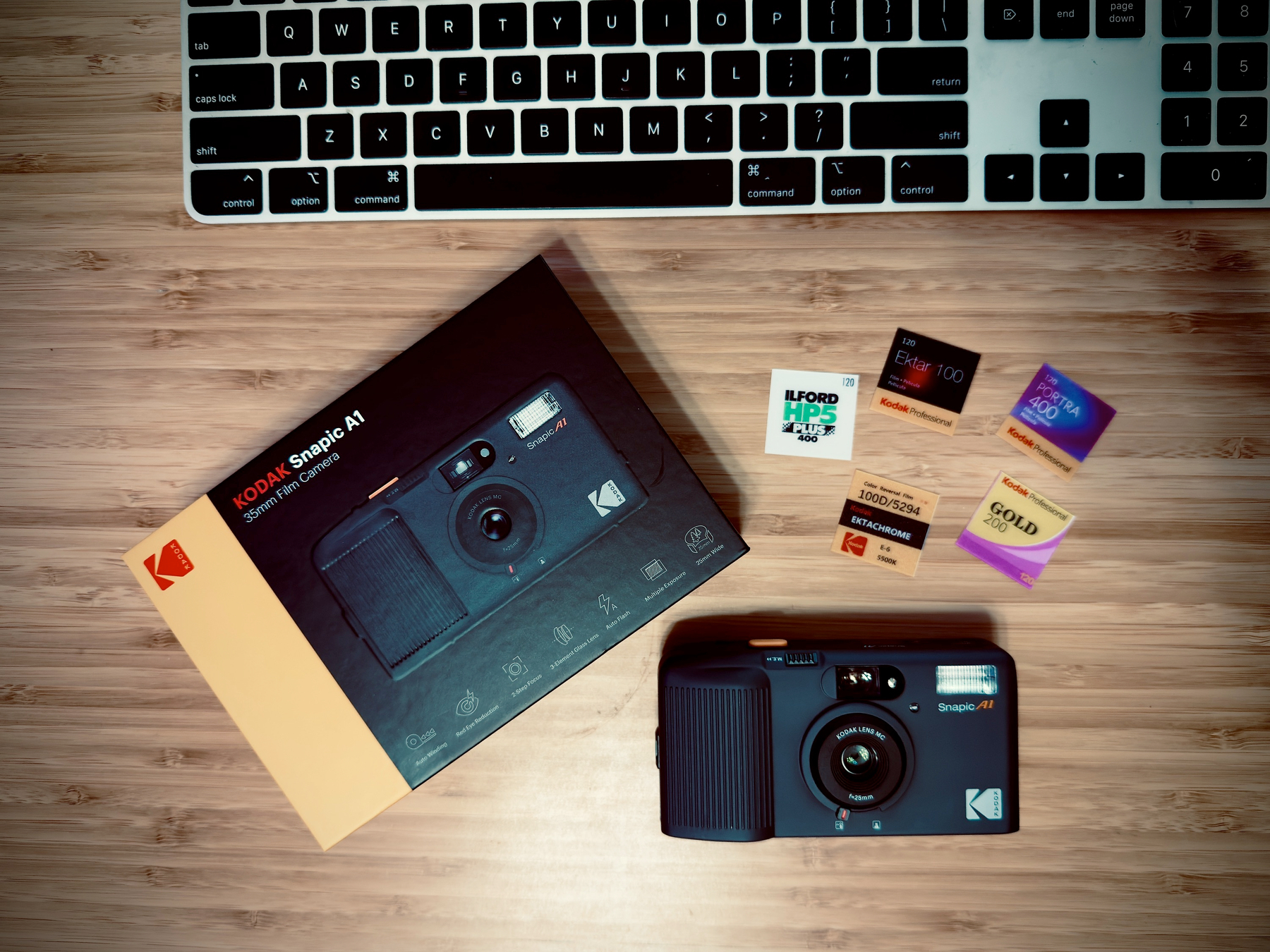 Auto-generated description: A Kodak Snapic Ai instant camera is placed on a wooden surface alongside its box, several film packs, and a part of a keyboard.