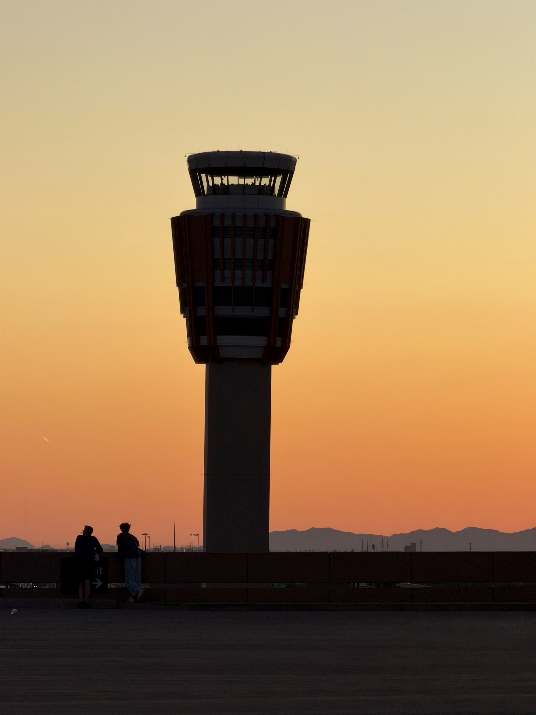 Auto-generated description: A control tower stands silhouetted against a sunset sky with two people walking in the foreground.
