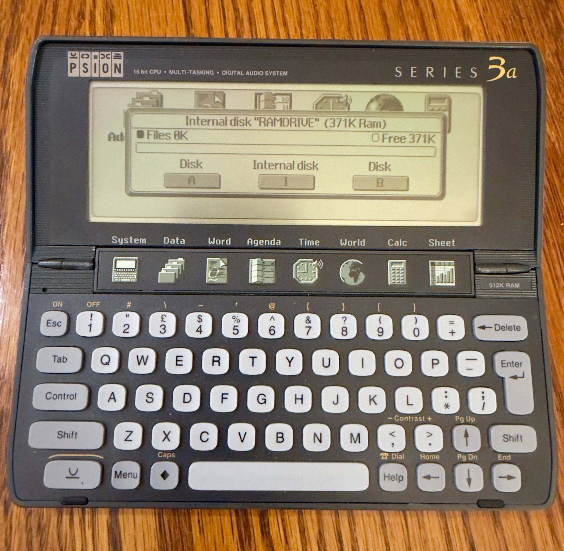 Auto-generated description: A vintage Psion Series 3a personal digital assistant with an open clamshell design and QWERTY keyboard rests on a wooden surface.