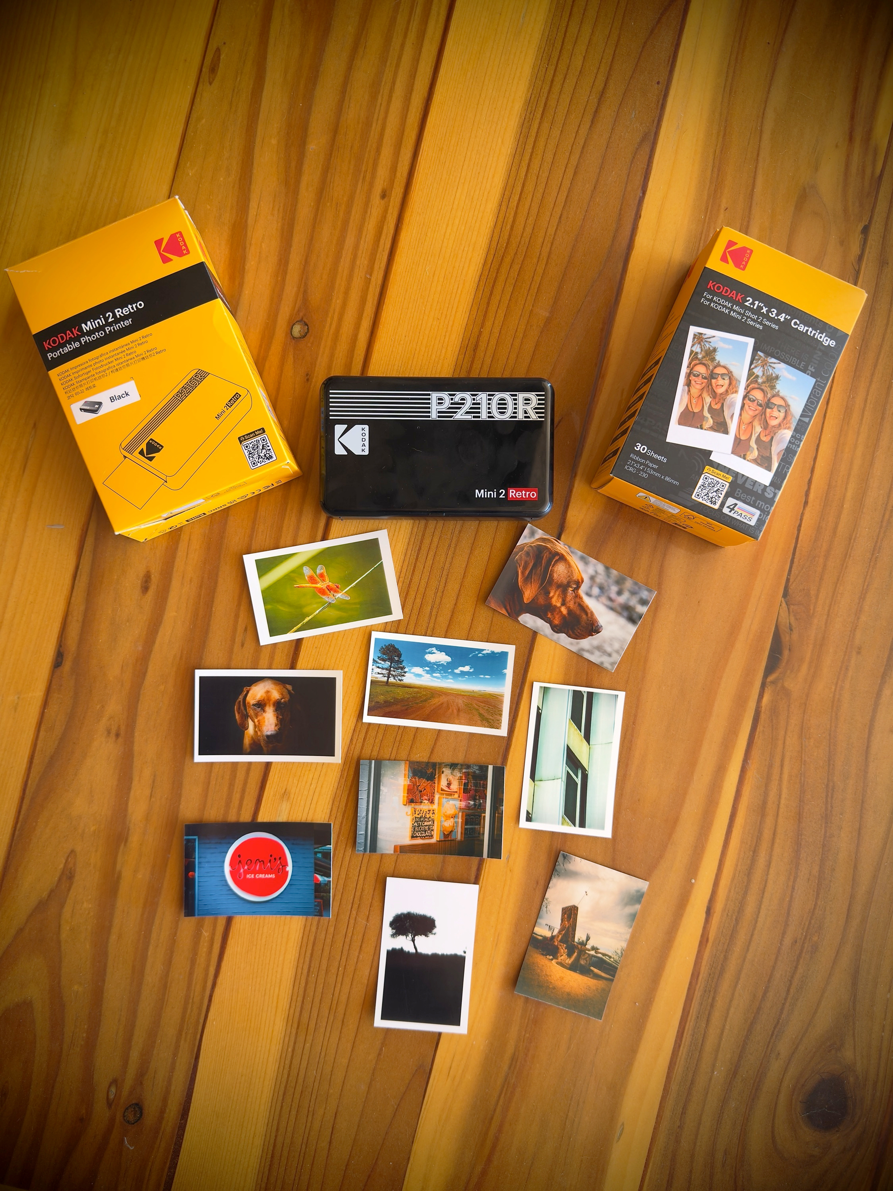 Auto-generated description: A Kodak mini camera and a selection of small printed photos are arranged on a wooden surface alongside two Kodak packaging boxes.