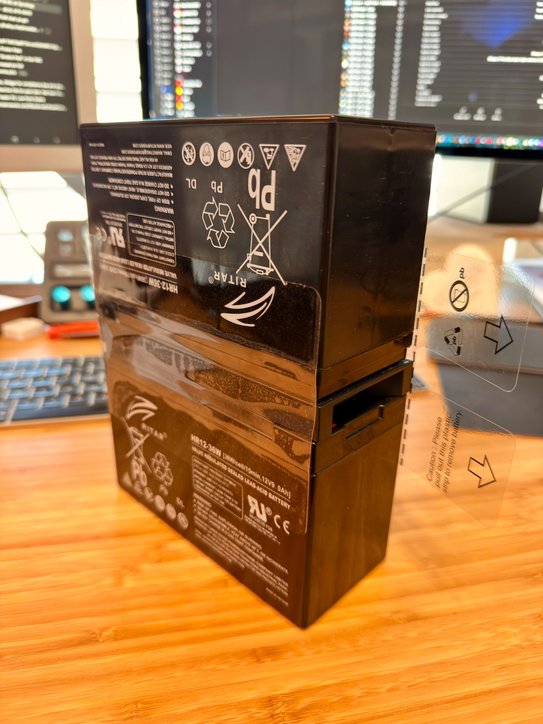 Auto-generated description: A black battery pack with various symbols and labels is positioned on a wooden surface near a computer keyboard and screen.
