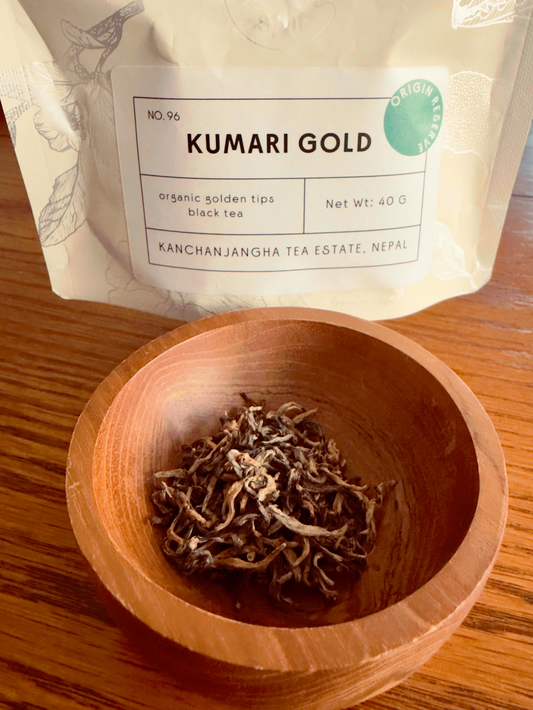 Auto-generated description: A wooden bowl filled with loose leaf tea is placed in front of a package labeled Kumari Gold from Kanchanjangha Tea Estate, Nepal.