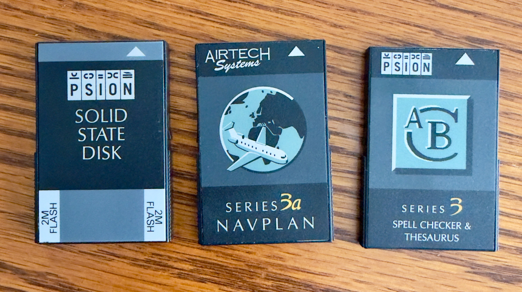 Auto-generated description: Three vintage Psion device modules are displayed on a wooden surface, featuring a solid state disk, a navigation planning system, and a spell checker & thesaurus.