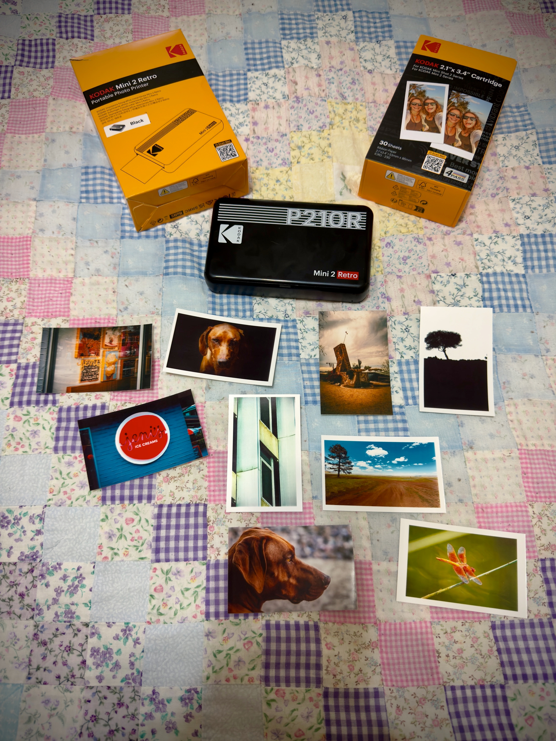 Auto-generated description: Various printed photos and two Kodak photo printer packages are arranged on a patchwork quilt background.