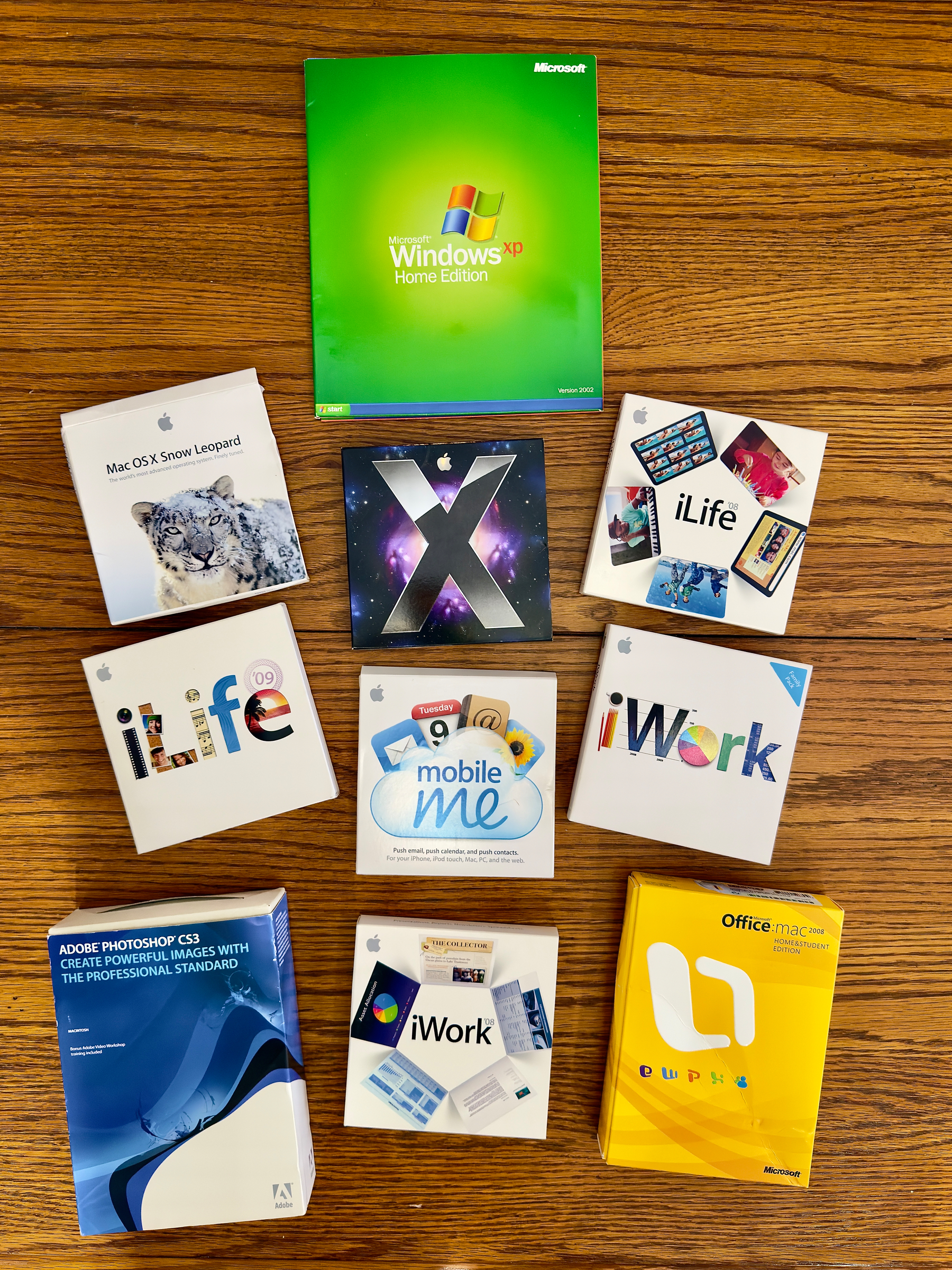 Auto-generated description: Various software packages for Windows XP, Mac OS X, iLife, iWork, MobileMe, Adobe Photoshop CS3, and Microsoft Office for Mac are arranged on a wooden surface.