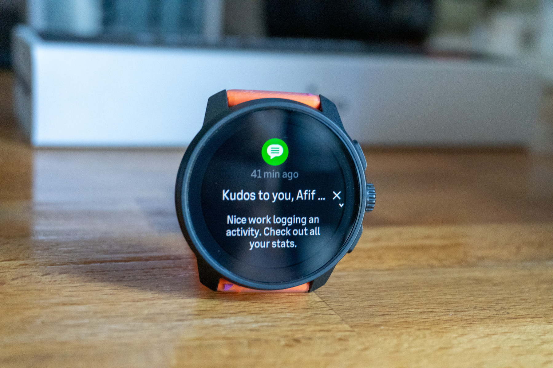A smartwatch with an orange strap displays a notification praising a user for logging activity and suggesting they check their stats.