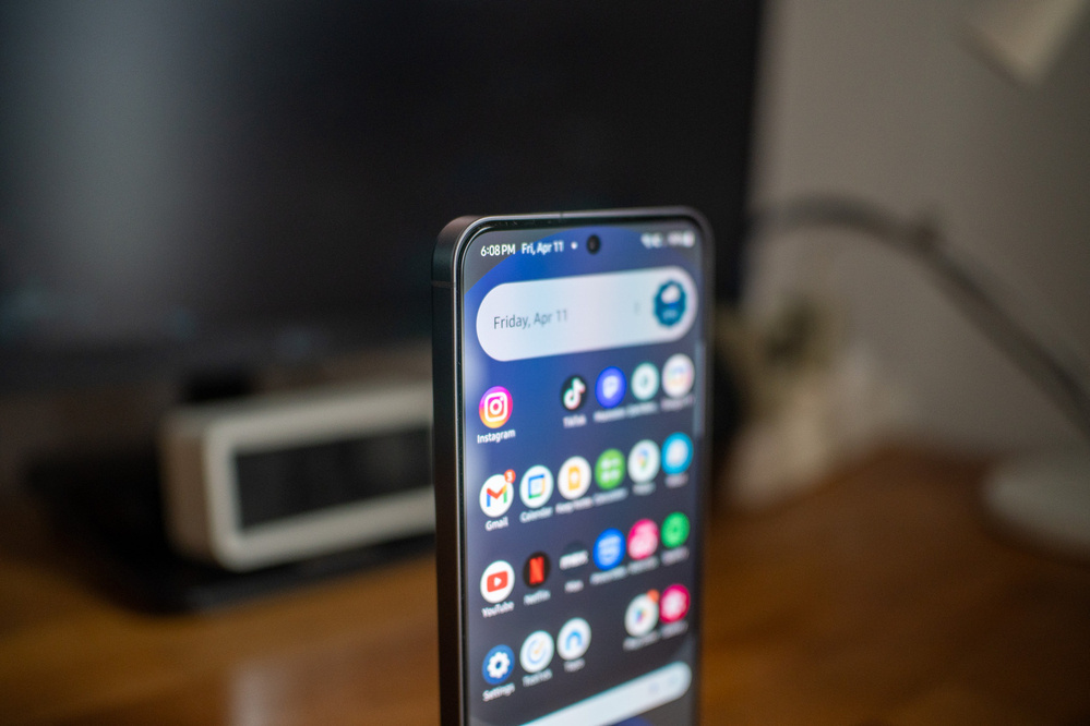 A smartphone displaying a variety of app icons is positioned upright on a wooden surface with a blurred background.