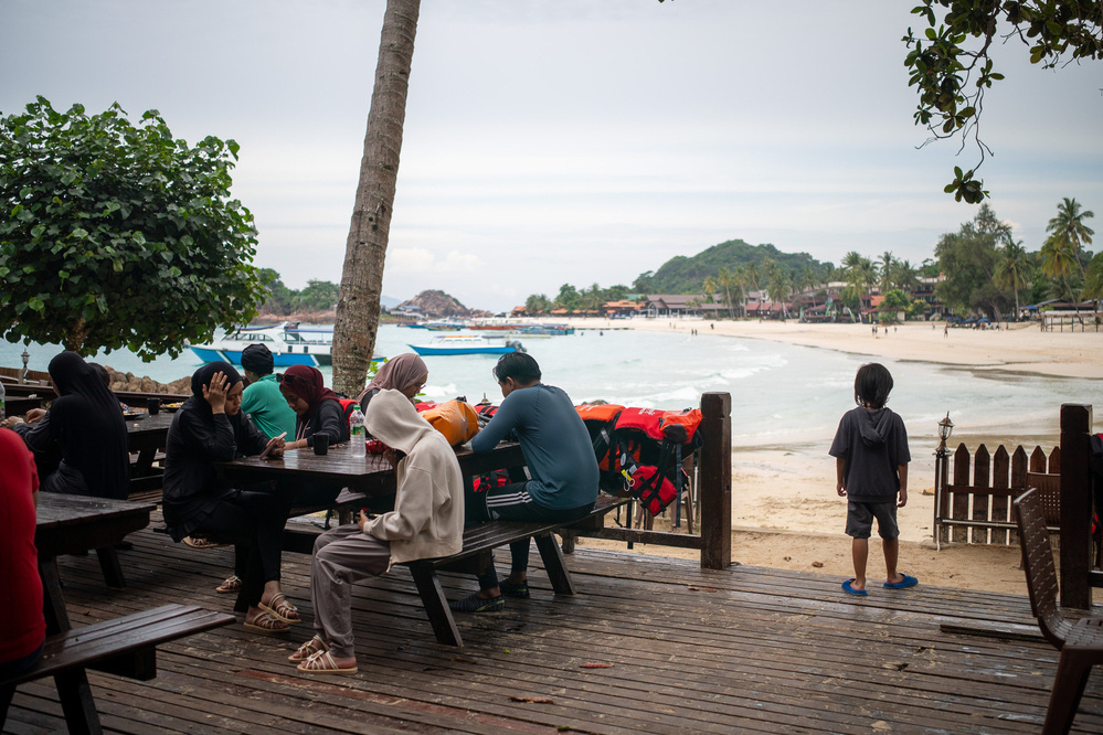People are sitting at outdoor tables overlooking a beach with boats and a hilly landscape in the background.