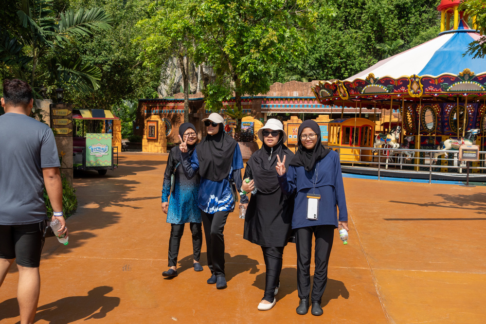 Four people are walking on an amusement park path, with a carousel and trees in the background.